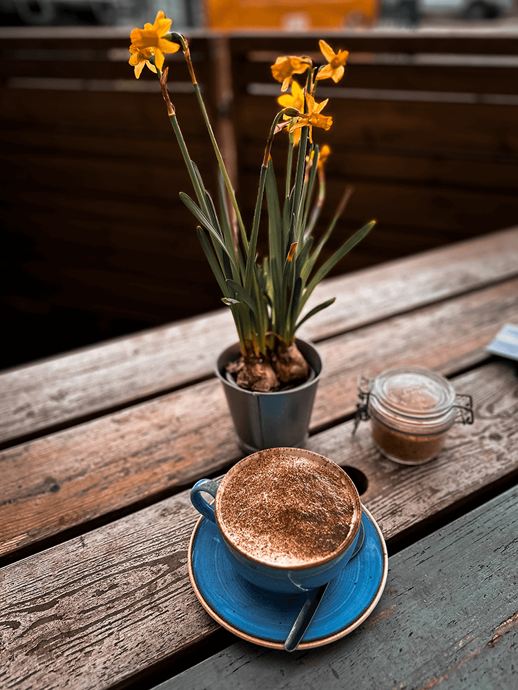 A blue mug of coffee next to some daffodils on a café bench.